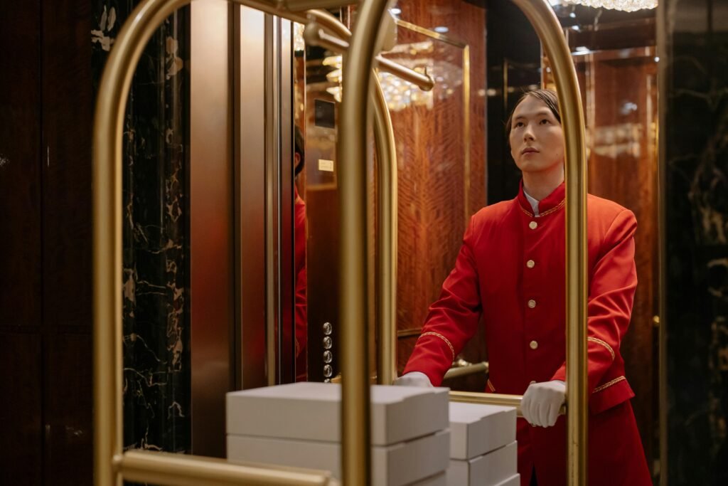 A hotel porter in a red uniform pushes a luggage cart in a luxurious elevator.
